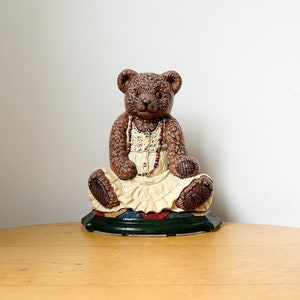 Könnte beinhalten: Ein brauner Teddybär, der ein weißes Kleid und eine Perlenkette trägt, sitzt auf einem bunten Teppich. Der Bär ist ein Türstopper.