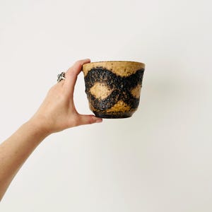 May include: A ceramic cup with a textured, speckled tan surface featuring a dark brown, rough-hewn horizontal band pattern that wraps around the exterior. The vessel has a wide, open rim and a slightly tapered base, showcasing a rustic, earthy aesthetic.