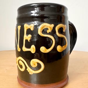 May include: A black ceramic beer mug with the word "GUINNESS" in raised, cream-colored lettering. The mug has a handle and a brown base. The mug is likely used for drinking beer.
