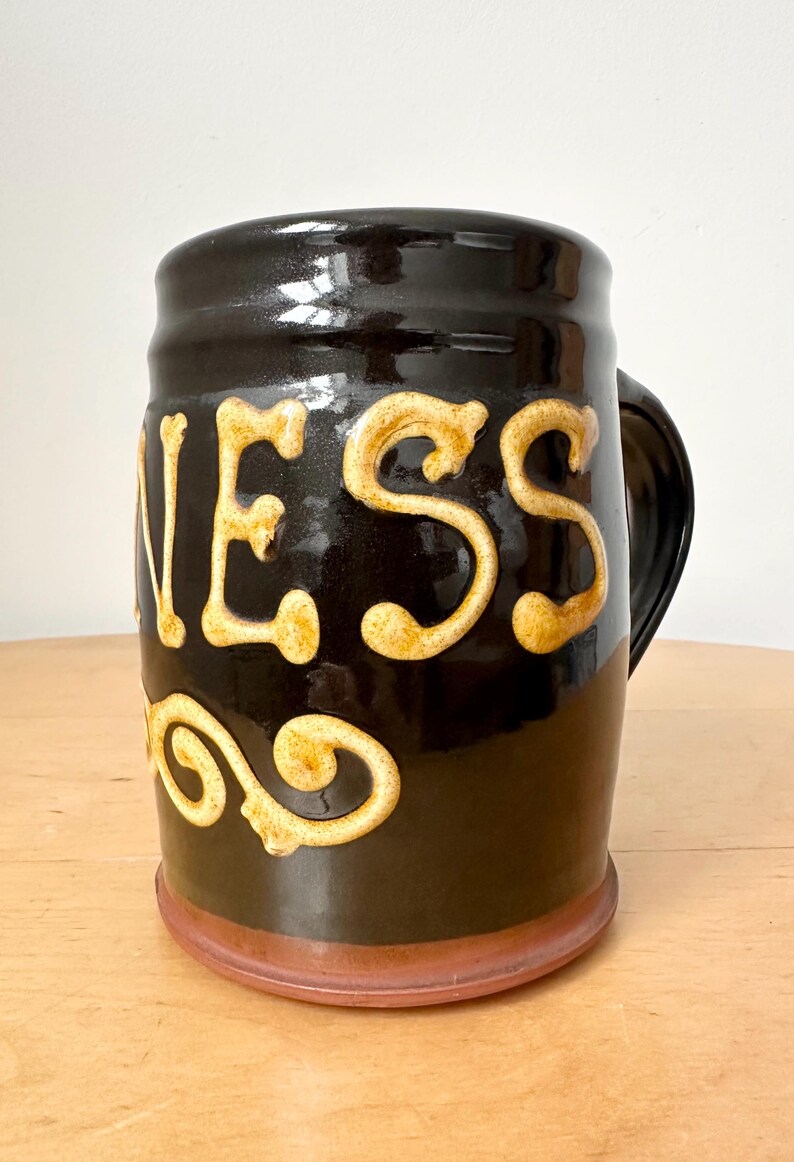 May include: A black ceramic beer mug with the word "GUINNESS" in raised, cream-colored lettering. The mug has a handle and a brown base. The mug is likely used for drinking beer.