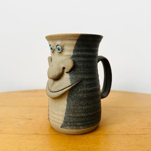 Può includere: Una tazza in ceramica con un volto scolpito caratterizzato da un naso grande e una bocca larga e sorridente. La tazza presenta un design bicolore con una finitura in argilla beige chiaro non smaltata sul davanti e uno smalto grigio antracite.
