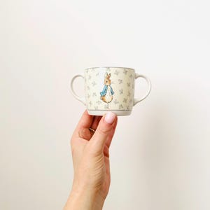 May include: A small, cream-coloured ceramic mug with two handles. The mug features a Peter Rabbit illustration and a pattern of small, light blue floral designs. The mug is held in a person's hand against a white background.