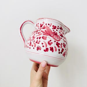 Puede incluir: Una jarra de cerámica blanca con un patrón pintado a mano de flores de color magenta y un pájaro estilizado. La jarra tiene un asa curva, un pico ancho y bandas horizontales de color magenta que rodean la base y el borde del recipiente.