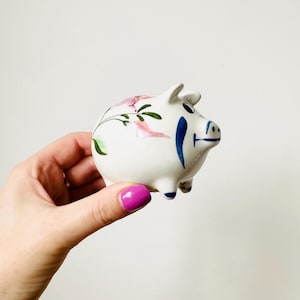 May include: A small, white ceramic piggy bank with hand-painted floral and abstract blue designs. The pig has a rounded body, small ears, and four tiny feet. The floral design includes pink and green accents.