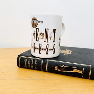 Puede incluir: Una taza de cerámica blanca con el texto "ORIENT EXPRESS" en letras marrones. La taza tiene un asa y está colocada sobre un libro oscuro con letras doradas. La taza tiene un diseño vintage.