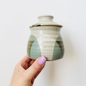 Puede incluir: Un pequeño tarro de cerámica hecho a mano con tapa. El tarro es cilíndrico con una base beige claro y un diseño geométrico verde y marrón. La tapa tiene un pequeño pomo. El tarro se sostiene en una mano.