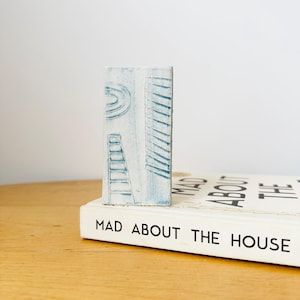 May include: A light blue ceramic rectangle with etched architectural designs, including a ladder and lines, sits on a book titled "MAD ABOUT THE HOUSE". The ceramic piece is a light blue colour.