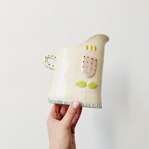 May include: Cream-colored ceramic pitcher with a whimsical floral design. The pitcher features a handle with black polka dots, a pink flower with black dots, and yellow accents. The base has a blue and green dotted border.