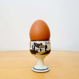 May include: A brown egg sits in a white and cream-colored egg cup. The egg cup has a painted scene of a house with the word "Portland" on it. The base of the egg cup has a blue stripe.