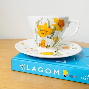 May include: A white teacup and saucer set featuring a floral design of yellow daffodils and green leaves. The teacup and saucer are placed on a blue book with the word "LAGOM" printed on the cover.