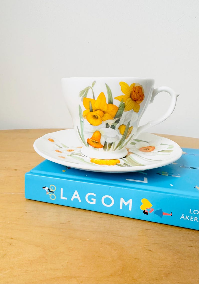 May include: A white teacup and saucer set featuring a floral design of yellow daffodils and green leaves. The teacup and saucer are placed on a blue book with the word "LAGOM" printed on the cover.