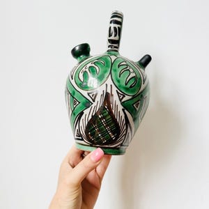 May include: A ceramic pitcher with a green, white, and black design. The pitcher has a handle, a spout, and two smaller spouts. The design includes geometric shapes and floral motifs. The pitcher is held by a hand against a white background.