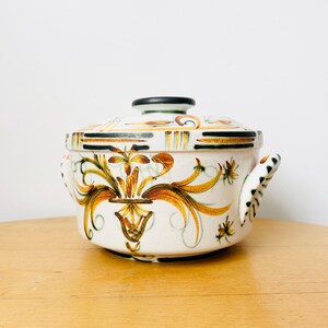 May include: A white ceramic casserole dish with a lid, featuring a floral design in shades of gold, brown, and black. The dish has two handles and a black knob on the lid. The design is symmetrical and detailed.