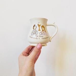Puede incluir: Taza de cerámica color crema con ribete dorado, con ilustraciones de dos niños y un cáliz. La taza tiene el texto "On Your First Holy Communion" y un asa curvada.