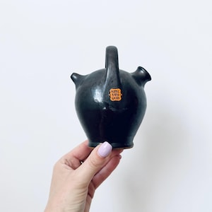 May include: A dark brown ceramic jug with a handle and two spouts. The jug has a rounded body and a small, orange and white label that reads "Kunst Hand Werk". The jug is held in a hand against a white background.