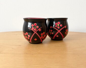 Puede incluir: Dos tazas de cerámica negra con diseños florales rojos y amarillos. Las tazas son redondas y tienen una base ancha.