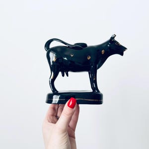 May include: A black ceramic cow figurine with gold accents. The cow is standing on a rectangular base, and the figurine has a glossy finish. The cow has gold spots and a curved tail. The figurine is held by a hand.