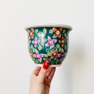 Op de afbeelding: Een teal keramische plantenpot met een bloemenontwerp met roze, oranje en groene bloemen. De pot is handgeschilderd met een witte rand.