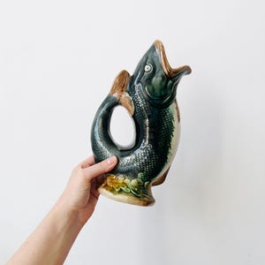 Może przedstawiać: Ceramiczny dzbanek w kształcie ryby z ciemnozielonym i czarnym łuskowatym korpusem, szeroko otwartym pyskiem pełniącym funkcję dzióbka oraz zakrzywionym ogonem tworzącym uchwyt. Podstawa ozdobiona jest wypukłymi żółtymi i zielonymi kwiatami.