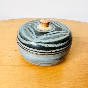 Op de afbeelding: Een ronde keramische pot met deksel, voorzien van een donkerblauw en zwart glazuur met een lichtblauw bladvormig patroon op het deksel. Het deksel heeft een kleine, centrale, lichtbruine knop. De basis van de pot heeft een lichtblauwe afwerking.