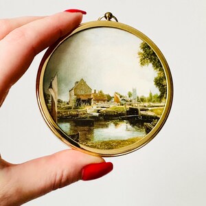 May include: A round, gold-toned pendant with a miniature landscape painting. The painting depicts a waterside scene with buildings, trees, and a church steeple. The pendant is held by a hand with red nail polish.