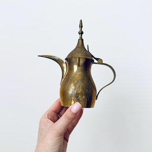 May include: A brass dallah coffee pot featuring a long, curved spout, a pointed lid with a finial, and a curved handle. The body of the pot is decorated with intricate, etched geometric patterns and traditional motifs across the metallic surface.
