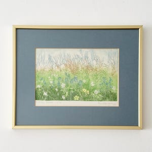 May include: A framed print featuring a landscape illustration of green grass, blue and white wildflowers, and bare trees in the background. The artwork is enclosed in a thin gold metal frame with a dark blue matting and includes handwritten text.