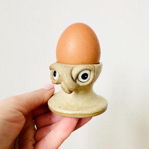 May include: A whimsical, beige ceramic egg cup with a face design, holding a brown egg. The egg cup has large, cartoonish eyes and a textured surface, adding a playful touch to the kitchen decor.