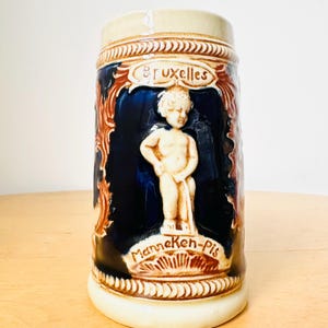 Op de afbeelding: Een keramische bierkan met een blauwe en bruine glazuur. De kan heeft een reli&euml;fbeeldhouwwerk van een kleine jongen die urineert. De kan is beschreven met de woorden "Manneken-Pis" en "Bruxelles".