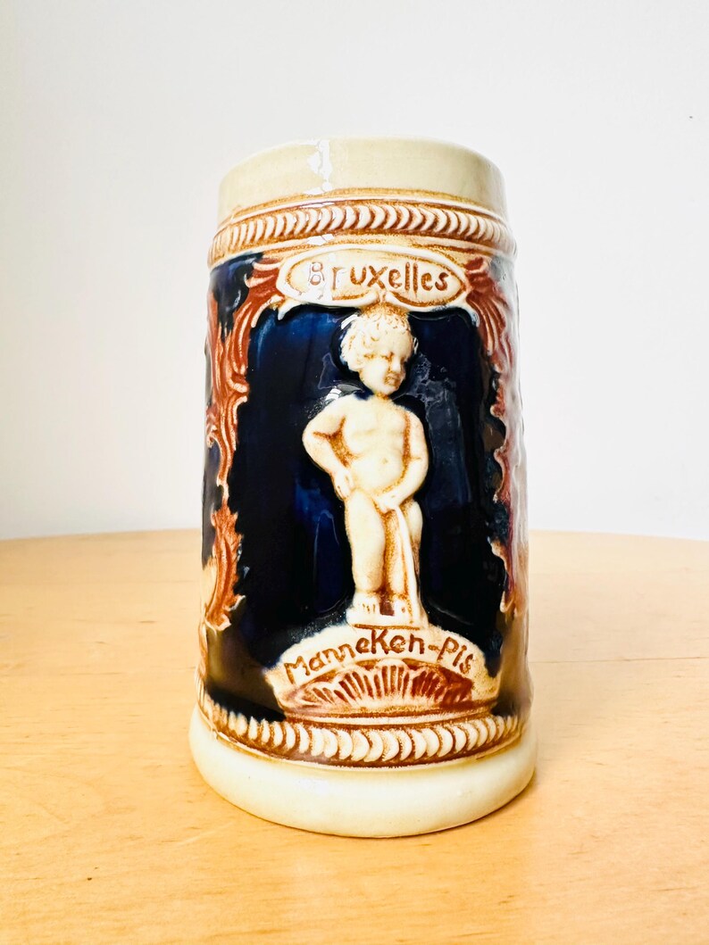 Op de afbeelding: Een keramische bierkan met een blauwe en bruine glazuur. De kan heeft een reli&euml;fbeeldhouwwerk van een kleine jongen die urineert. De kan is beschreven met de woorden "Manneken-Pis" en "Bruxelles".