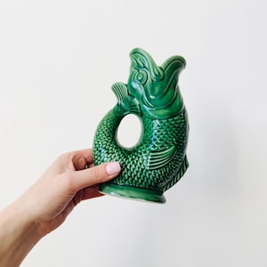 Może przedstawiać: Zielony ceramiczny dzban w kształcie ryby z otwartymi ustami i uchwytem. Dzban ma teksturowany, przypominający łuski wzór i małą płetwę. Dzban jest trzymany na białym tle.