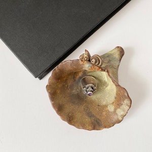 K&ouml;nnte beinhalten: Ein Keramik-Ringhalter in Form einer Blume mit einer gr&uuml;n-braunen Glasur. Ein silberner Ring mit einem violetten Edelstein liegt im Ringhalter.