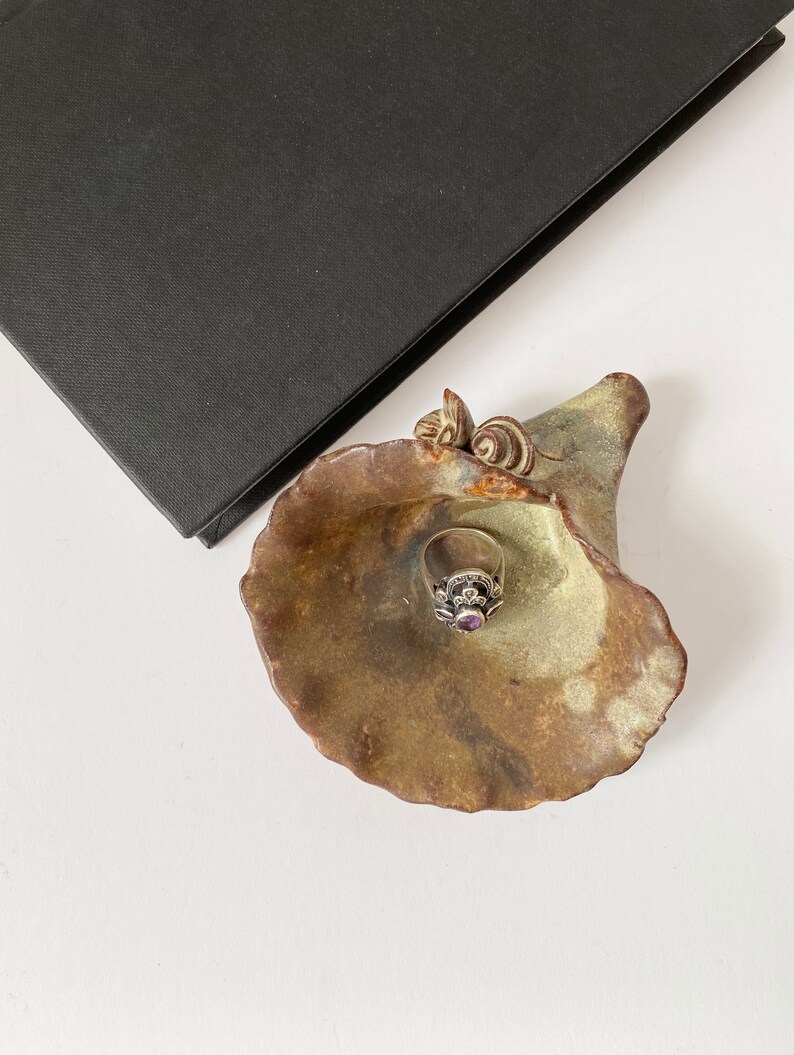 K&ouml;nnte beinhalten: Ein Keramik-Ringhalter in Form einer Blume mit einer gr&uuml;n-braunen Glasur. Ein silberner Ring mit einem violetten Edelstein liegt im Ringhalter.