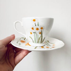 May include: White teacup and saucer set featuring a floral design of white daffodils with yellow centers and green stems. The cup has a curved handle and sits on a matching saucer. The set is held by a hand.