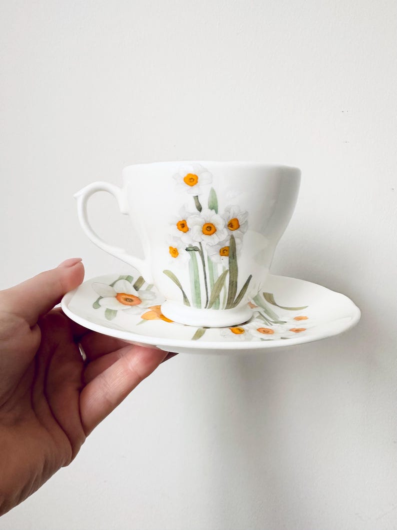 May include: White teacup and saucer set featuring a floral design of white daffodils with yellow centers and green stems. The cup has a curved handle and sits on a matching saucer. The set is held by a hand.