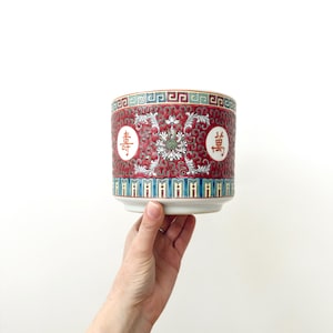 May include: A cylindrical ceramic planter featuring a vibrant red background with intricate white floral scrollwork, teal and gold geometric borders, and two white circular medallions containing black Chinese characters on the front surface.
