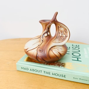 May include: A brown and cream ceramic vase with a rounded body and a single handle. The surface features embossed eucalyptus leaves, seed pods, and a relief scene of a person riding a horse. The vase has a smooth, matte finish with gradient tones.