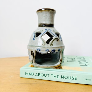 May include: A ceramic candle holder in grey and brown. It features diamond-shaped cut-outs and blue decorative patterns. The candle holder is placed on a book titled "MAD ABOUT THE HOUSE".