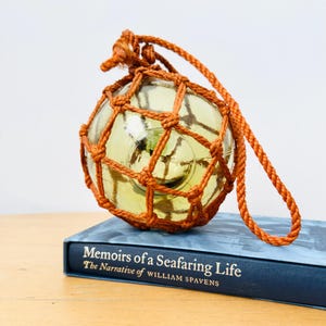 May include: A clear glass float with a yellow tint, encased in a woven orange net, and a matching rope handle. The float rests on a blue book titled "Memoirs of a Seafaring Life".
