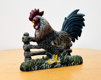 Op de afbeelding: Een gietijzeren haanfiguur met een rode kam en lellen, een bruin en grijs gevederd lichaam en een donkerblauwe staart. De haan staat op een groene basis naast een hekwerk, met felgele voeten die zichtbaar zijn in het gras.