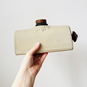 Può includere: Una borsa dell'acqua calda beige vintage con tappo e manico marroni. La bottiglia rettangolare ha una superficie strutturata e un motivo floreale decorativo attorno al tappo. Un oggetto classico per il comfort e il calore.
