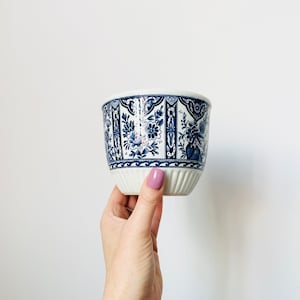 May include: A white ceramic planter with a blue floral pattern. The planter has a slightly flared shape and a ribbed base. The blue design features stylized flowers and foliage. The planter is held by a hand against a white background.