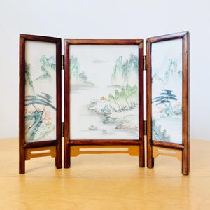 May include: A small, three-panel folding screen featuring a landscape painting. The screen has a dark wood frame and a light background with a watercolour-style painting of mountains, trees, and water. It is hinged and stands on a wooden surface.