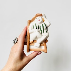 May include: A ceramic figurine depicting a figure wearing a brown hat and a white swimsuit with vertical green stripes, seated on a brown chair. The figure has a hand raised to their head and is rendered in a glossy, painted finish with defined facial features.