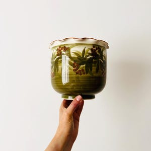 May include: A ceramic planter with a scalloped rim and a glossy finish. The exterior features a hand-painted design of green leaves and small red berries against a dark olive green base with horizontal brushstroke textures throughout the body.