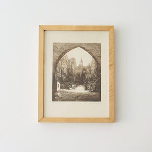 Puede incluir: Impresión en tonos sepia enmarcada de una escena de calle europea, vista a través de un arco. La impresión presenta elementos arquitectónicos detallados y una sensación de profundidad, encerrada en un marco de madera clara. La obra evoca una estética vintage.