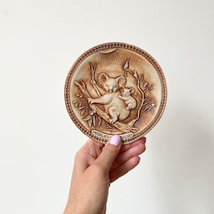 May include: A brown ceramic plate with a raised image of a koala and joey, surrounded by eucalyptus leaves and birds. The plate has a decorative border and the words "KOALAS - AUSTRALIA" written on the bottom.