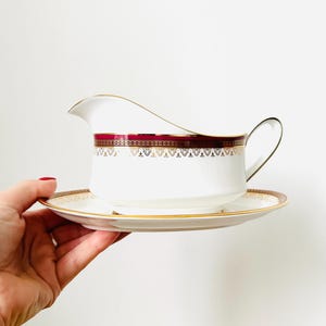 Puede incluir: Juego de salsera y platillo blancos con un borde decorativo burdeos y dorado. La salsera tiene un asa y un pico. El platillo es redondo y tiene un borde a juego. El conjunto está sostenido por una mano.