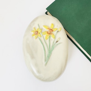 Puede incluir: Una caja de cerámica ovalada blanca con un diseño en relieve de tres narcisos amarillos con tallos y hojas verdes.