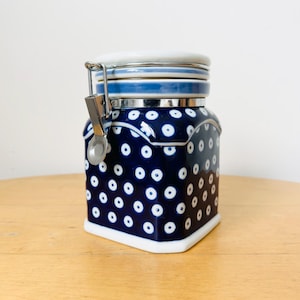 May include: A ceramic jar with a lid and metal clasp. The jar is navy blue with white polka dots and a white base. The lid is white with a blue band. A small metal spoon is attached to the clasp.